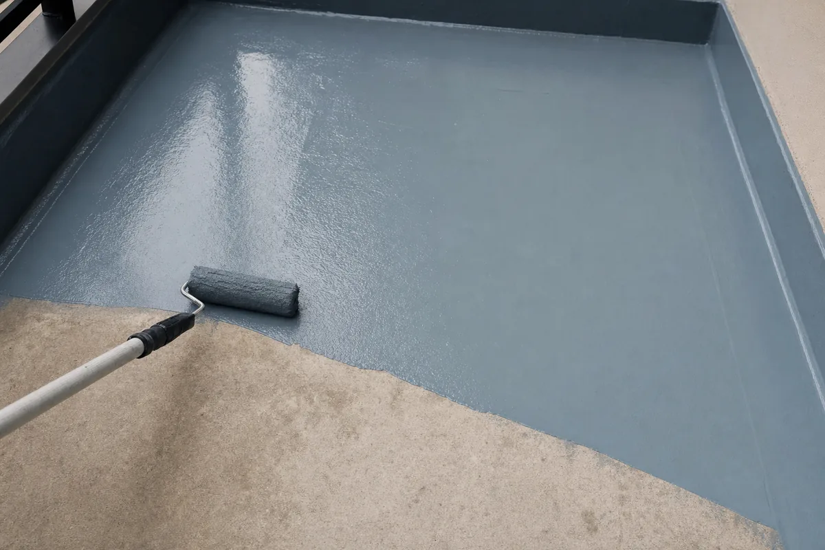 Grey-blue liquid membrane being rolled across a bare concrete balcony slab, a wet edge halfway across and the wall upstand already coated — illustrative example